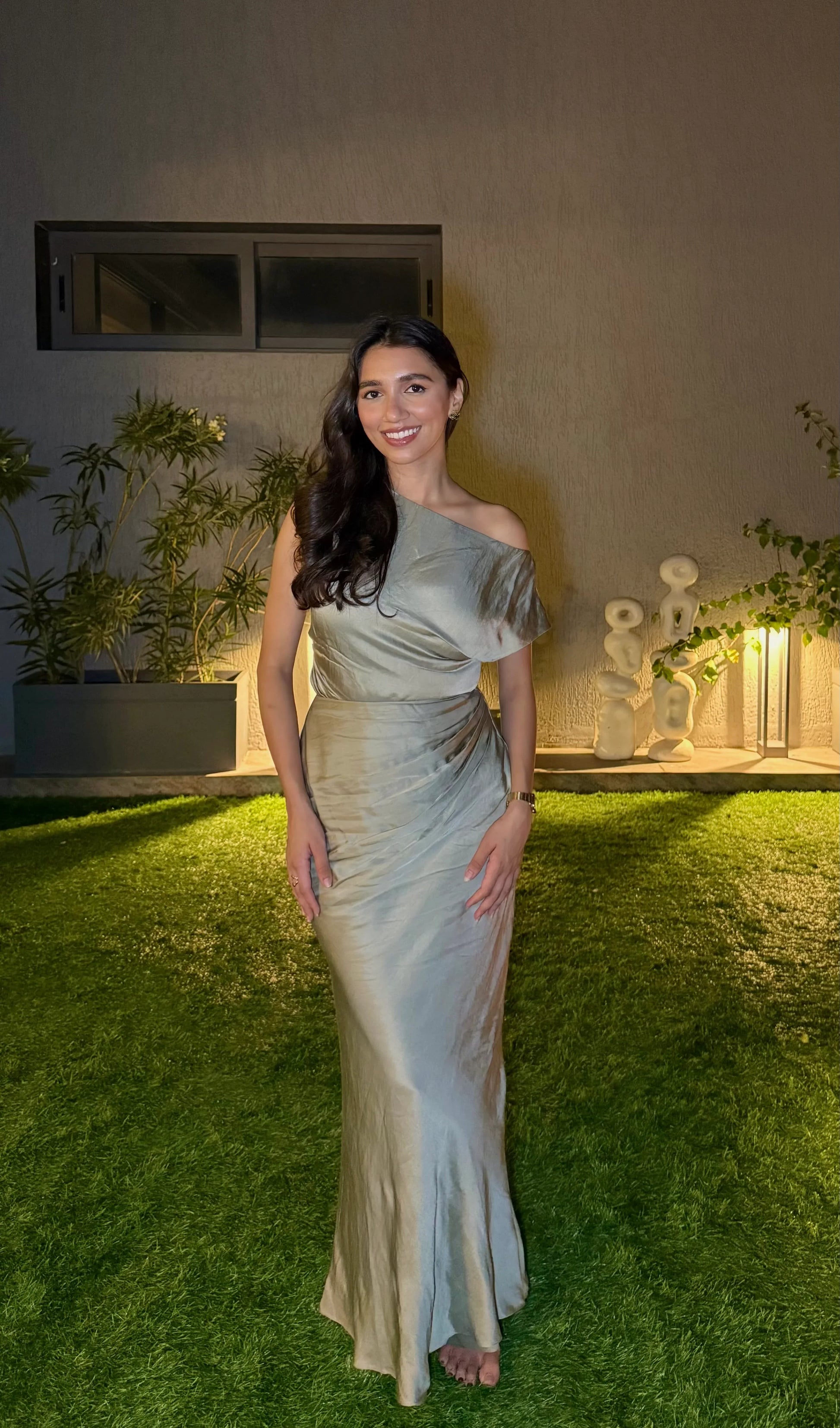 Woman in sage green off-shoulder satin top and maxi standing on grass at night in a landscaped garden